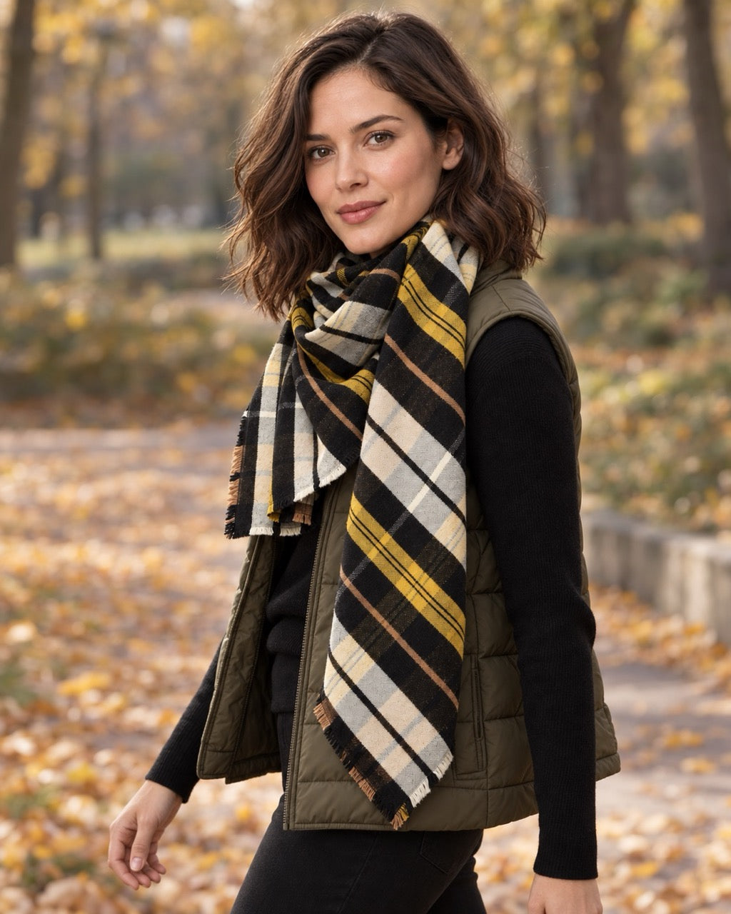 Model wearing an s.Oliver black and mustard checked scarf over a black sweater and olive quilted vest in an autumn park.