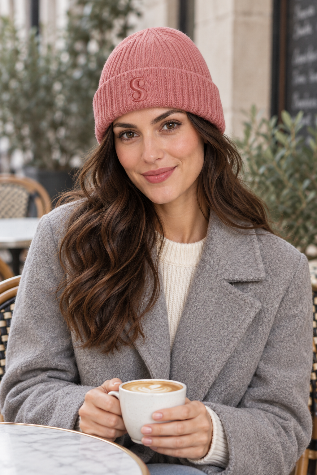 Woman wearing a dusty pink s.Oliver knitted beanie and a grey coat at an outdoor cafe.